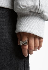 Inel argintiu cu un design decupat, purtat pe o mână. Hanorac gri cu manșete ribbed și blugi închis în fundal, fără detalii texturate vizibile.