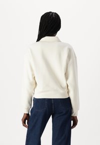 Felpa bianca corta con colletto, polsini e orlo a coste, abbinata a jeans blu scuro. Sul retro presenta un design liscio e senza decorazioni.