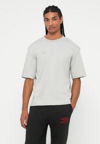 Hombre con cabello trenzado vestido con una camiseta ligera gris de Umbro y pantalones negros con el logo rojo de Umbro, de pie frente a un fondo blanco.