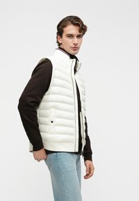 Jaunas vyras, dėvintis baltą siūtu švarką ant tamsios ilgomis rankovėmis marškinio ir šviesiai mėlynų džinsų, stovi prieš paprastą baltą foną.