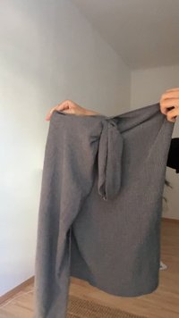 Mani che tengono una gonna in tessuto grigio con un dettaglio a nodo contro uno sfondo di muro semplice e pavimento in legno.
