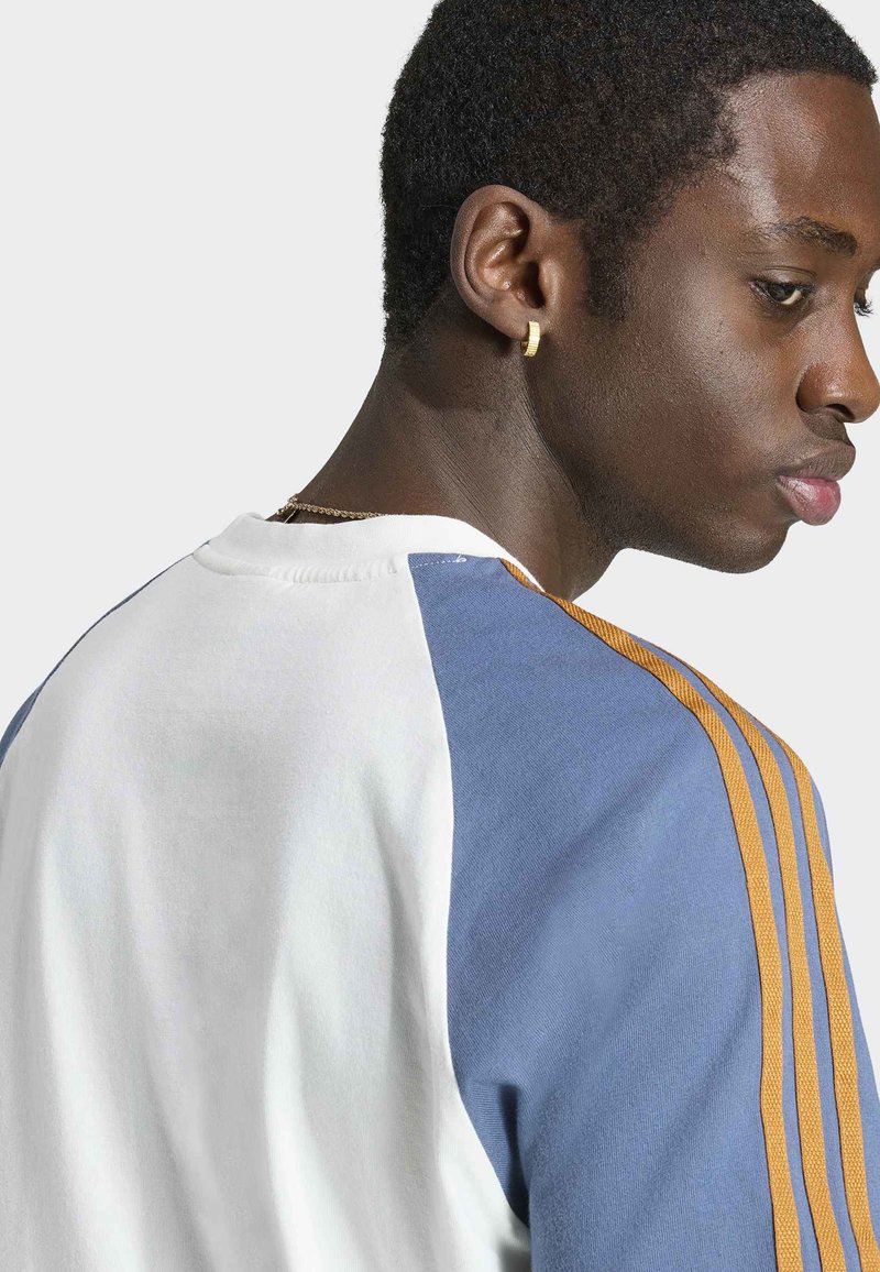 Junger Mann mit kurzen Haaren, der ein weißes und blaues Hemd mit orangefarbenen Streifen trägt, seitlich betrachtet, mit einem goldenen Creolenohrring im linken Ohr.