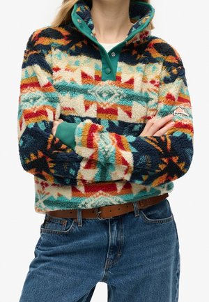 Femme portant un pull polaire à motifs multicolores avec un col vert à boutons-pression, une ceinture marron et un jean bleu, les bras croisés sur la poitrine.