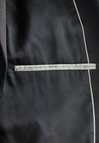 Tessuto nero con un sottile motivo diagonale, rifinito con un bordo grigio lungo il contorno e una tasca orizzontale con cuciture grigie.