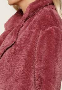 Cappotto in finta pelliccia di un rosa intenso con colletto, morbido al tatto e dalla forma leggermente over-size. Dettagli delle cuciture visibili lungo i lati.