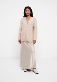 Cardigan mohair bej deschis, cu decolteu adânc în V, deschidere cu nasturi și croială lejeră, asortat cu o fustă maxi bej dintr-un material mătăsos și pantofi slip-on.