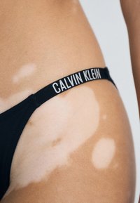 Črn Calvin Klein tanga, ki je narejena iz mehkega materiala, ima ožji pas z logotipom in gladke robove, ki se lepo prilegajo različnim tonom kože.
