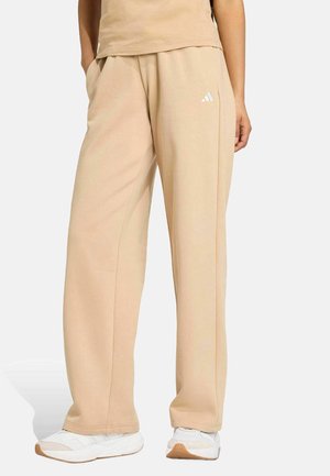 Personne portant un pantalon de survêtement large beige avec le logo Adidas et des baskets blanches, debout les mains dans les poches devant un fond uni.