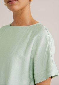 Ljusgrön kortärmad topp med en avslappnad passform, som har en bred halsringning och en mjuk, något strukturerad tygkvalitet. Sömdetaljer är synliga.