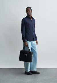 Tamsiai mėlyna ilgomis rankovėmis marškiniai, laisvas siluetas, šviesiai mėlynos plačios kojos džinsai, juoda odinė portfelis su priekiniais užtrauktukais, juodi batai, stovintis prieš neutralų foną.