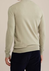 Pull vert clair en maille avec poignets et ourlet côtelés. Vue de dos montrant une texture lisse et une coupe ajustée. Associé à un pantalon bleu marine foncé.