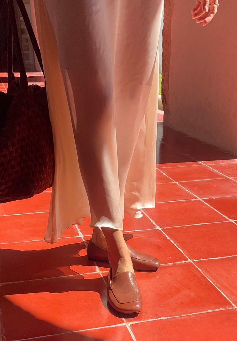 Jambes en robe légère beige clair et mocassins en cuir marron debout sur un sol carrelé rouge avec la lumière du soleil projetant des ombres. Main avec bague visible.