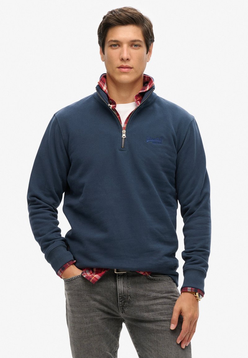 Pullover in pile blu navy con colletto a zip. Indossato sopra una camicia a quadri rossi, con polsini a costina e un piccolo logo ricamato sul petto.