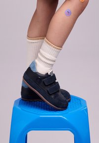 Sneaker blu navy con una combinazione di suede e pelle, dotati di due strap in velcro e accenti blu chiaro, appoggiati su uno sgabello blu.