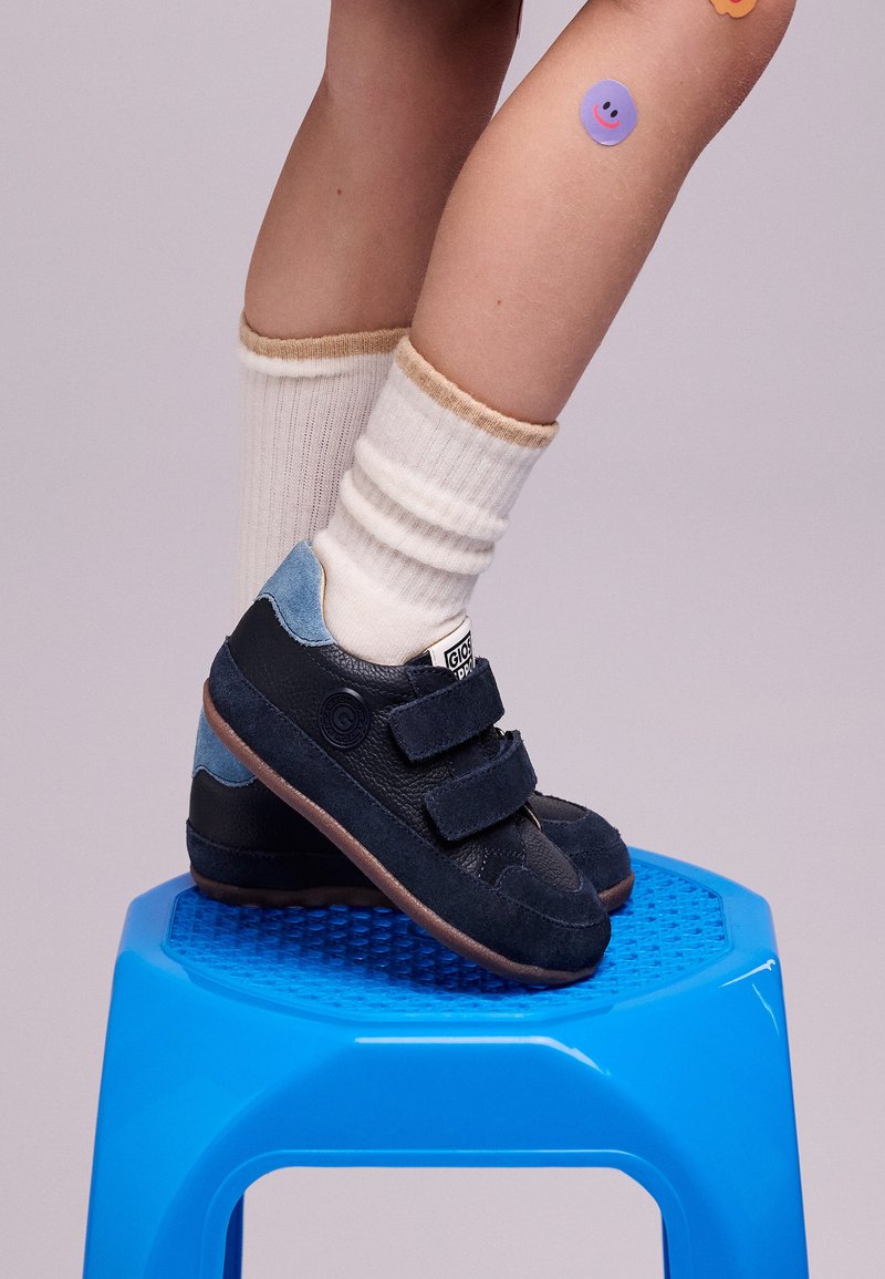 Sneaker blu navy con una combinazione di suede e pelle, dotati di due strap in velcro e accenti blu chiaro, appoggiati su uno sgabello blu.