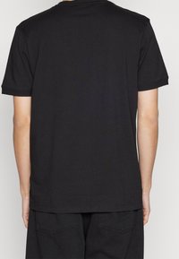 Persona che indossa una semplice t-shirt nera a maniche corte e pantaloni neri, vista di spalle su uno sfondo chiaro e uniforme.