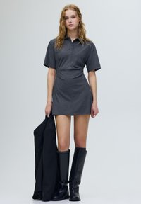 Szürke galléros ruha rövid ujjakkal, testhezálló derékkal és gombos előrésszel, térd feletti fekete csizmával és fekete dzsekivel párosítva.