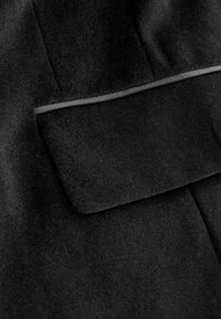 Tissu noir avec une texture lisse et douce, doté d'un détail de poche. Fini avec une fine ligne de matériau contrastant.