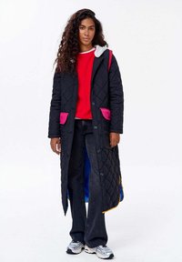 Žena s dlouhými kudrnatými vlasy, oblečená v dlouhém černém prošívaném kabátu s růžovými kapsami, červeném svetru, tmavých džínách a stříbrných teniskách, stojící.