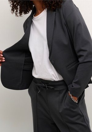 Person iført mørkegrå blazer åbnet for at vise indvendig lomme, hvid t-shirt, matchende bukser med snor og sølvkædearmbånd på håndleddet.