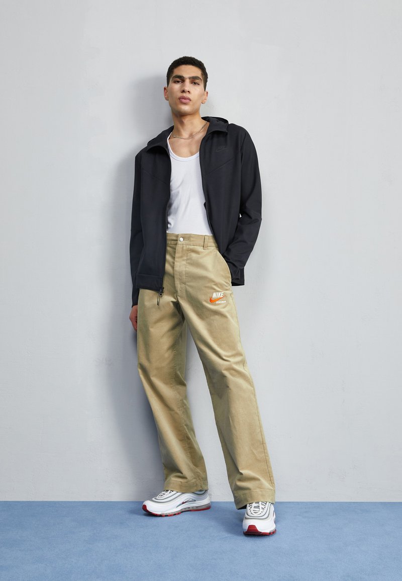 Giacca nera con zip sopra una canotta bianca, abbinata a pantaloni beige con un logo Nike arancione e sneakers bianche con dettagli rossi.