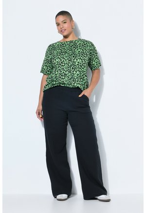 Mujer con cabello corto vestida con una camisa verde de estampado de leopardo, pantalones anchos negros, zapatos blancos y pendientes de aro dorados, posando con confianza.