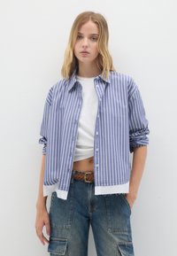 Camicia a righe blu e bianche con maniche arrotolate, indossata sopra un top corto bianco. Abbinata a pantaloni cargo in denim a vita alta.