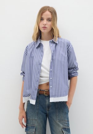 Camisa de botones a rayas azul y blanca con mangas enrolladas, llevada sobre un top corto blanco. Combinada con pantalones cargo de mezclilla de cintura alta.