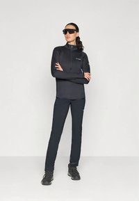 Γυναίκα που φοράει μαύρο εξωτερικό μπουφάν, παντελόνι, ορειβατικά μποτάκια και μεγάλα μαύρα γυαλιά ηλίου, στέκεται με τα χέρια σταυρωμένα μπροστά σε απλό φόντο.