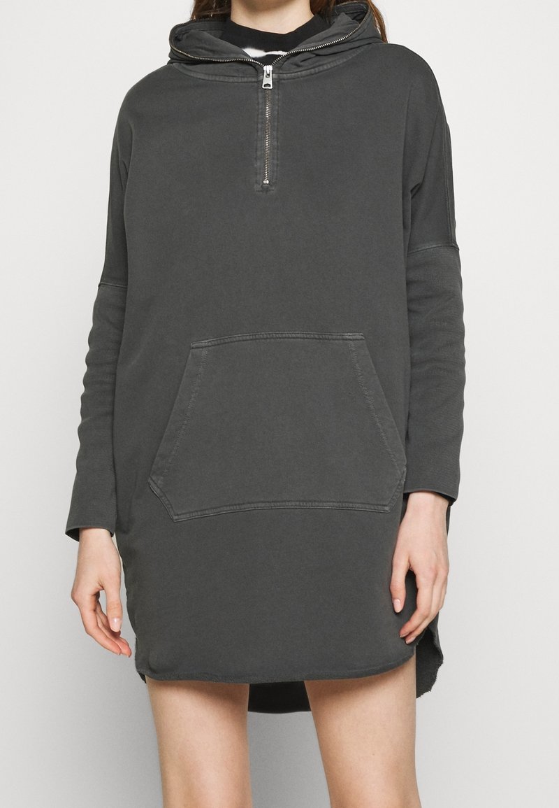 Femme portant une robe à capuche gris foncé à manches longues avec une fermeture éclair sur le devant et une poche kangourou, debout devant un fond clair uni.