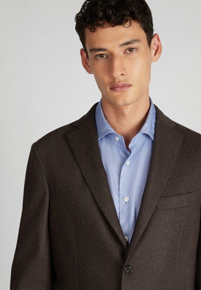 Blazer in lana marrone con spalle strutturate, chiusura a un bottone e taschino sul petto, indossato sopra una camicia azzurro chiaro con una trama liscia.