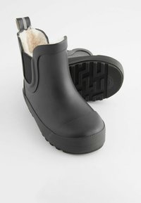 Bottes de cheville en caoutchouc noir avec une douce doublure en fourrure crème, semelle extérieure texturée et panneaux latéraux élastiques pour un enfilage facile. Design minimaliste.