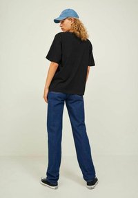 Black oversized T-shirt featuring short sleeves, paired with blue denim jeans and black and white sneakers. Light blue baseball cap worn backwards.