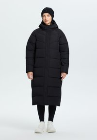 Abrigo acolchado largo negro con capucha, que presenta un diseño acolchado, cremallera frontal completa y puños acanalados. Combinado con leggins negros y zapatillas blancas.
