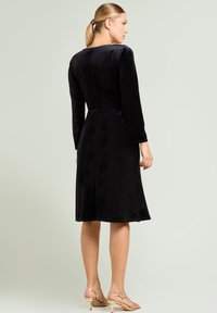 Schwarzes Samtkleid mit langen Ärmeln, ausgestelltem Saum und rundem Ausschnitt. Glatte Textur und minimalistisches Design. Kombiniert mit goldfarbenen Riemchenabsätzen.