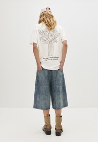 Person med langt bølget hår iført en hvid t-shirt med et floralt vase-design, lyse denim korte bukser, brune støvler og en pink kasket, vendt væk.