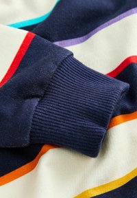 Marineblaue Rippbündchen auf einem gestreiften Stoff mit horizontalen Linien in Creme, Rot, Orange, Türkis und Lila; glatte Textur.