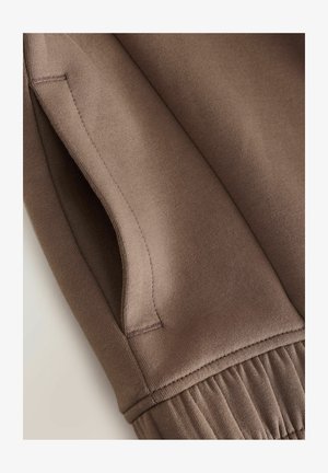 Tela de sudadera marrón con textura suave, que cuenta con un bolsillo lateral, dobladillo acanalado y un detalle sutil de costuras.