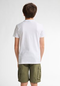 Camiseta de algodón blanca con mangas cortas y cuello redondo, combinada con pantalones cortos tipo cargo en color verde oliva, que cuentan con bolsillos laterales y un acabado texturizado.