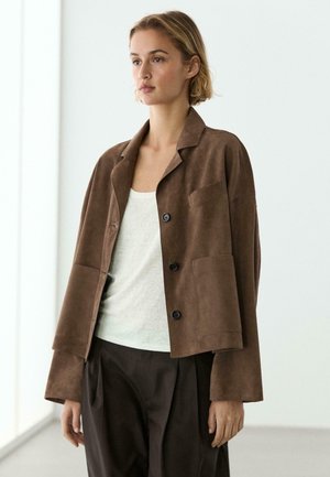 Jonge vrouw met kort blond haar, die een bruine suède jas, wit topje en donker losse broek draagt, staand in een lichte minimalistische ruimte.