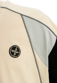 Camiseta deportiva beige y negra con detalles grises, un tejido texturizado y un logo circular bordado en el lado izquierdo.