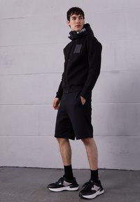 Černá mikina s kapucí na zip a náprsní kapsou, doplněná černými kraťasy. Model nosí černé tenisky s bílými podrážkami a ladící černé ponožky.