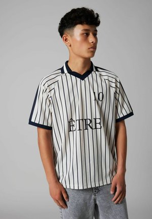 Joven con cabello rizado que lleva una camisa blanca de rayas finas con cuello negro y el texto "ÊTRE 10", de pie frente a un fondo gris liso.