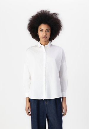 White button-up shirt with a relaxed fit, long sleeves, and a pointed collar. Fabric appears smooth and lightweight, paired with dark denim pants.