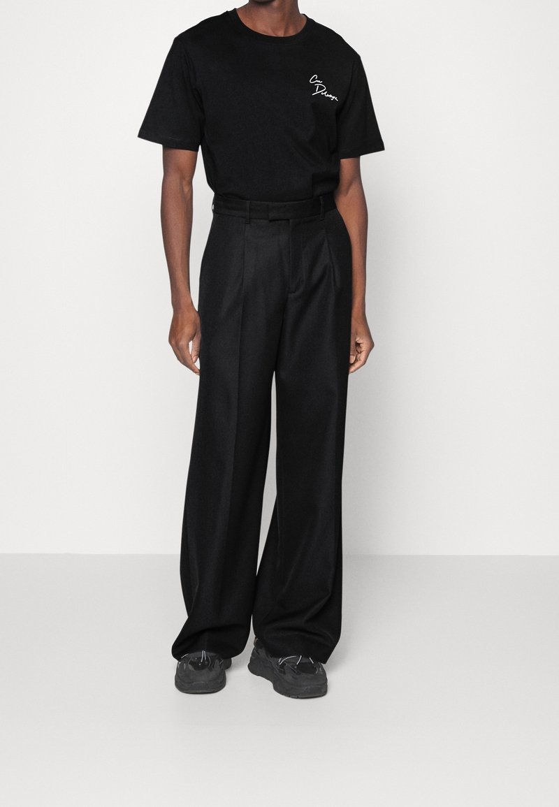 Černé bavlněné tričko s bílým logem na hrudi, v kombinaci s volnými černými kalhotami a černými teniskami s jemnými akcenty.