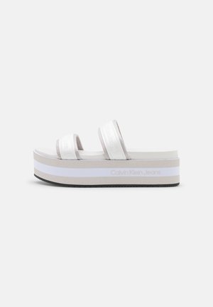 Witte plateau-slidesandaal met twee stoffen bandjes en "Calvin Klein Jeans"-branding op de zijkant van de zool, tegen een effen achtergrond.