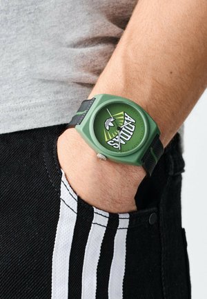 Montre en plastique verte avec un cadran rond arborant un motif en éclat de soleil et le texte "ADIDAS". Bracelet ajustable en tissu noir tissé avec des accents verts.