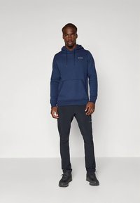 Marinblå hoodie med framficka och justerbar huva; kombinerad med svarta byxor och stadiga svarta kängor, med en logotyp på bröstet.