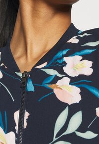 Detalj av en persons nacke och en marinblå zip-up-jacka med blommönster i rosa och blå blommor samt gröna löv.