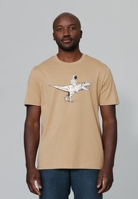 Lichtbruine t-shirt met korte mouwen met een dinosaurustekening en een astronaut die erop rijdt. Gemaakt van katoen, heeft een ronde halslijn en een rechte zoom.